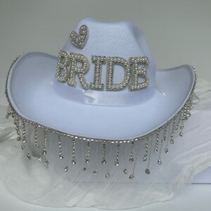 May include: White cowboy hat with a rhinestone trim and the word "BRIDE" spelled out in rhinestones. The hat also has a small rhinestone heart on the front.