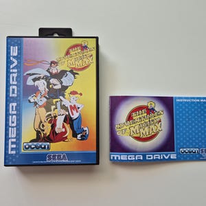 Op de afbeelding: Een Mega Drive videogamebox en instructiehandleiding voor "The Adventures of Mighty Max". De doos toont stripfiguren en de titel van het spel. De handleiding heeft een blauw stippenpatroon.