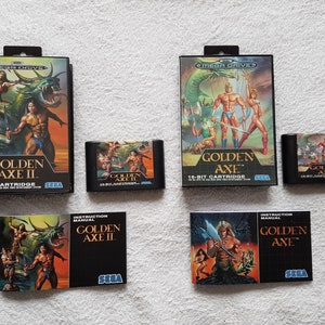 以下が含まれることがあります： ゴールデンアックスとゴールデンアックスIIのセガジェネシスビデオゲームカートリッジと説明書のコレクション。カートリッジは黒地に金色のアクセントが施され、金色の斧を持った戦士のイラストが描かれています。説明書は黒地に金色の文字で、カートリッジと同じイラストが描かれています。