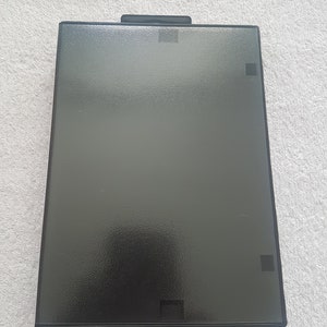 Puede incluir: Una caja de pl&aacute;stico negra con una superficie texturizada. La caja tiene una peque&ntilde;a abertura rectangular en la parte superior e inferior.