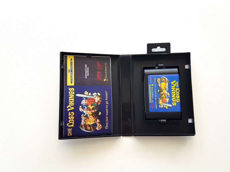 Puede incluir: Una caja de cartucho de juego Sega Genesis negra con un cartucho de juego azul y amarillo en su interior. El cartucho de juego es para el juego "The Lost Vikings". La caja tiene una carcasa de pl&aacute;stico negro con una ventana de pl&aacute;stico transparente para mostrar el cartucho de juego. El cartucho de juego tiene un fondo azul con texto amarillo e im&aacute;genes de los personajes del juego.