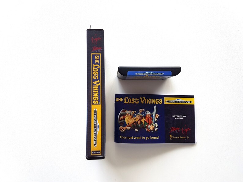 Puede incluir: Un cartucho de juego Sega Mega Drive y un manual de instrucciones para el juego "The Lost Vikings". El cartucho de juego es azul y negro con el texto "Sega Mega Drive" en &eacute;l. El manual de instrucciones es azul y negro con una imagen de los personajes del juego. El texto "The Lost Vikings" est&aacute; impreso en amarillo en el manual. El texto "They just want to go home!" est&aacute; impreso en blanco en el manual.