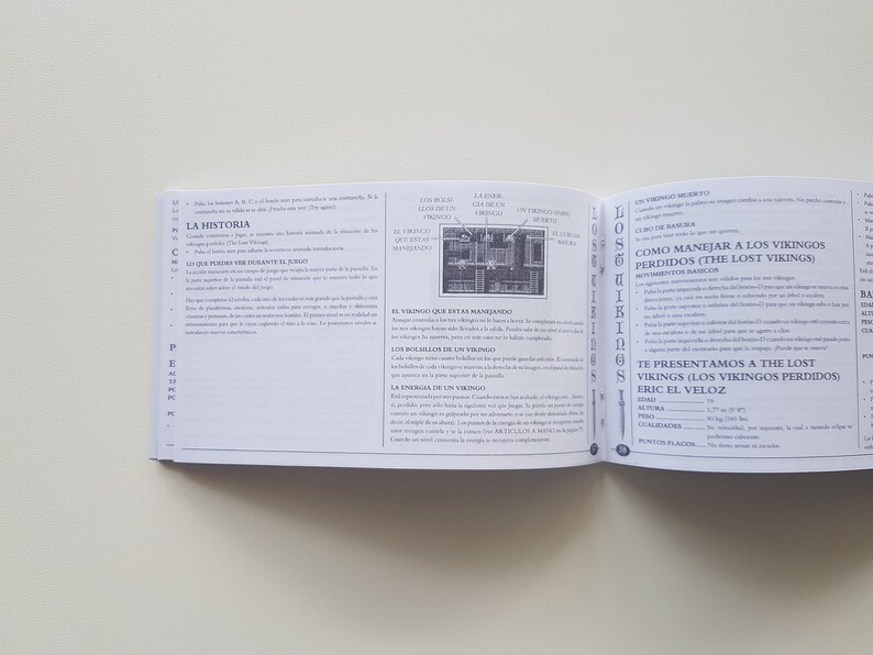 Puede incluir: Un libro abierto en una p&aacute;gina con el t&iacute;tulo "The Lost Vikings" y una descripci&oacute;n de c&oacute;mo jugar al juego. El libro es parte de un juego de mesa.