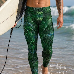Może przedstawiać: Zielone legginsy we wzór łuski. Osoba trzyma deskę surfingową na plaży. Osoba idzie w wodzie. Tło obejmuje ocean, fale i słoneczne niebo.