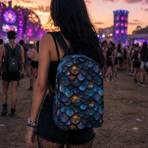 May include: A backpack with a blue, teal, and gold scale pattern. The backpack is worn by a person in a crowd at an outdoor event. The background features a sunset and large structures with bright lights. The backpack has black straps.