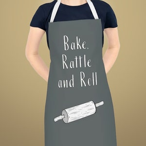Pode incluir: Um avental cinza com texto branco que diz "Bake. Rattle and Roll." Um rolo de massa branco é ilustrado no avental.