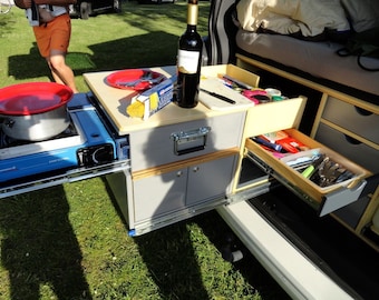 May include: A compact, portable kitchen setup for a van or camper. It features a pull-out counter with a stovetop, a sink, and storage drawers. The counter is made of wood and has a light gray finish. The drawers are made of wood and have a light brown finish. A bottle of wine is on the counter.