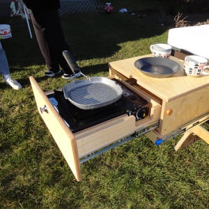 Puede incluir: Un cajón de madera con una estufa integrada y una sartén para asar. El cajón está abierto y la estufa es visible. Hay dos tazas y un plato en el mostrador sobre el cajón.