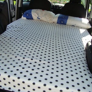 Op de afbeelding: Een wit beddengoed met zwarte stippen is in de achterkant van een auto gelegd, waardoor een geïmproviseerd bed ontstaat.