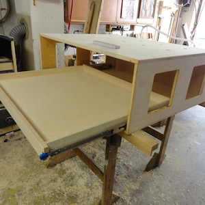 Può includere: Un banco da lavoro in legno color chiaro con un cassetto estraibile e due ritagli rettangolari. Il banco da lavoro è sostenuto da un telaio in legno e si trova in un'officina.