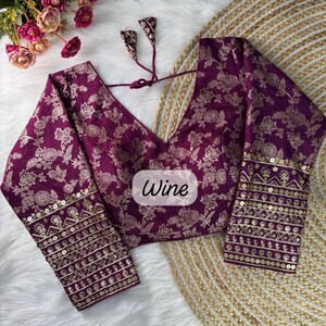 May include: A deep purple blouse featuring a gold floral pattern, long sleeves with gold sequin and embroidery detailing at the cuffs, a V-neckline, and a back tie closure. The center of the garment displays a label with the text Wine.