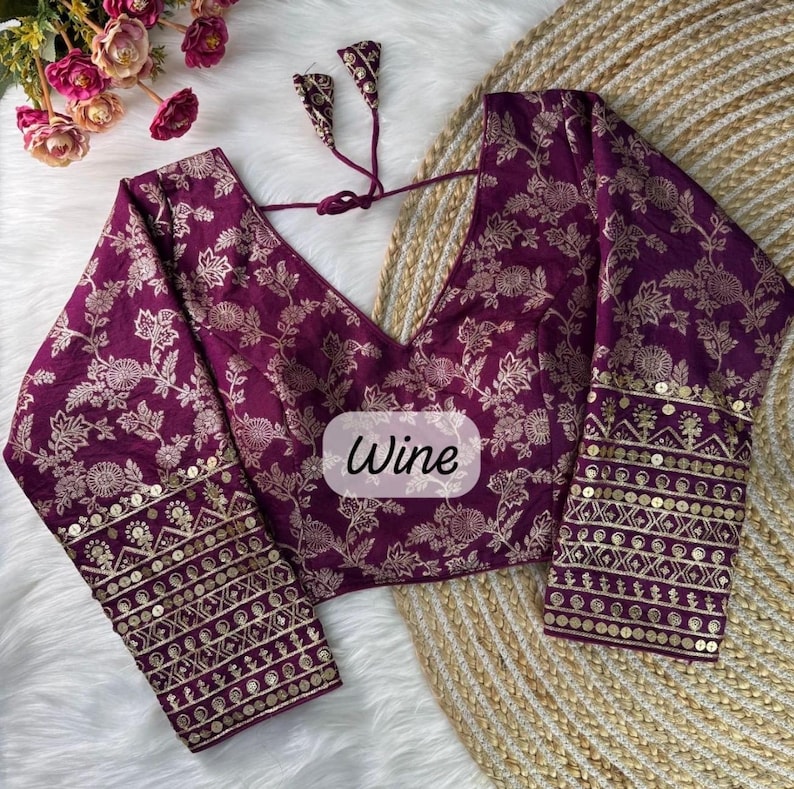 May include: A deep purple blouse featuring a gold floral pattern, long sleeves with gold sequin and embroidery detailing at the cuffs, a V-neckline, and a back tie closure. The center of the garment displays a label with the text Wine.