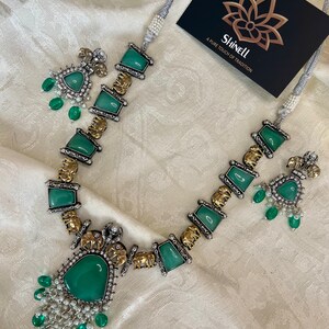 Op de afbeelding: Een zilveren en gouden ketting met groene edelsteen hangers en olifant bedels. De ketting heeft een bijpassend paar oorbellen met groene edelstenen.
