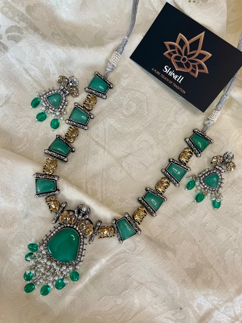 Op de afbeelding: Een zilveren en gouden ketting met groene edelsteen hangers en olifant bedels. De ketting heeft een bijpassend paar oorbellen met groene edelstenen.