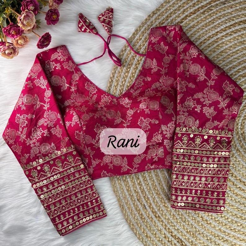 May include: A magenta long-sleeve blouse featuring a repeating gold floral pattern, a V-neckline with thin tie strings, and wide cuffs adorned with horizontal bands of gold embroidery and sequins. The word Rani is printed in the center of the blouse.