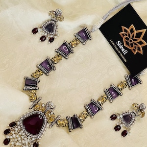 Op de afbeelding: Een zilveren en gouden ketting met een hanger van paarse steen en olifantbedels. De ketting wordt geleverd met bijpassende oorbellen met paarse stenen en rode kralen.