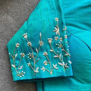 May include: A vibrant teal blouse featuring short sleeves adorned with intricate hand-embroidered floral patterns. The embroidery consists of delicate stems and small blossoms crafted from metallic gold and silver thread, creating a textured botanical design across the fabric surface.
