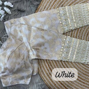 May include: A white blouse featuring gold floral embroidery throughout the fabric. The garment has long sleeves with detailed gold sequin embellishments and geometric patterns at the cuffs. The word White is printed in black text on the lower right.