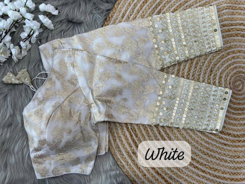 May include: A white blouse featuring gold floral embroidery throughout the fabric. The garment has long sleeves with detailed gold sequin embellishments and geometric patterns at the cuffs. The word White is printed in black text on the lower right.