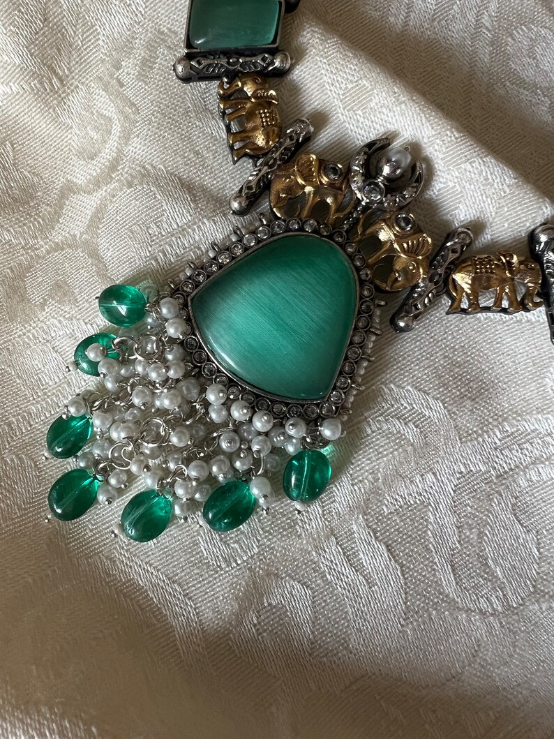 Op de afbeelding: Een zilveren en gouden ketting met een groene stenen hanger en een franje van groene en witte kralen.