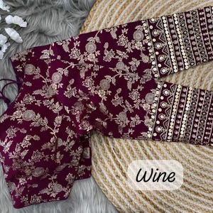 May include: A wine-colored blouse featuring a gold floral pattern throughout the fabric. The garment has three-quarter length sleeves with wide cuffs decorated with gold embroidery and circular sequins. The word Wine is printed in black text.