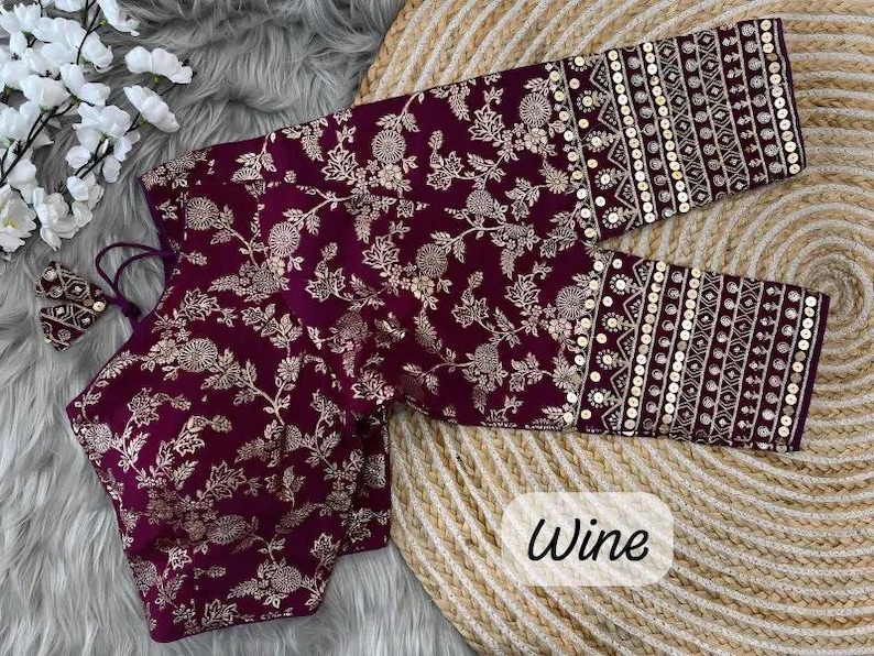 May include: A wine-colored blouse featuring a gold floral pattern throughout the fabric. The garment has three-quarter length sleeves with wide cuffs decorated with gold embroidery and circular sequins. The word Wine is printed in black text.