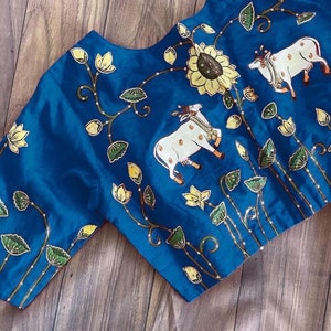 May include: A blue silk blouse with a floral and cow design. The blouse has a round neckline and short sleeves. The design features white cows, yellow sunflowers, green leaves, and yellow flowers.