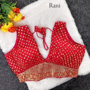 May include: A red sleeveless top with a deep V-neck and gold floral pattern. The bottom edge features a gold embroidered border. The top is displayed on a white fluffy surface. The word "Rani" is in the top right corner.