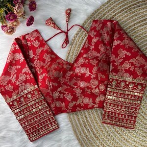 May include: A vibrant red blouse featuring a repeating gold floral pattern, a deep V-neckline, and long sleeves. The cuffs are embellished with horizontal bands of gold embroidery and sequins. Two thin red fabric ties with tassels are attached at the back.