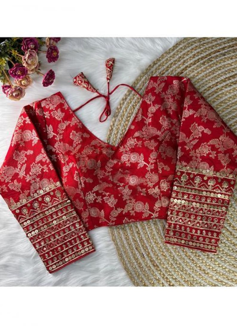 May include: A vibrant red blouse featuring a repeating gold floral pattern, a deep V-neckline, and long sleeves. The cuffs are embellished with horizontal bands of gold embroidery and sequins. Two thin red fabric ties with tassels are attached at the back.