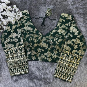 May include: A dark green blouse featuring a gold floral pattern throughout. The garment has a V-neckline with black tie strings at the back. The long sleeves are adorned with horizontal bands of gold sequins and intricate geometric embroidery.