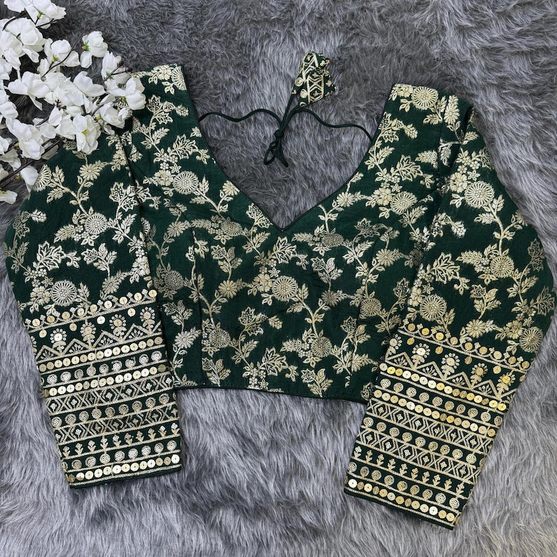 May include: A dark green blouse featuring a gold floral pattern throughout. The garment has a V-neckline with black tie strings at the back. The long sleeves are adorned with horizontal bands of gold sequins and intricate geometric embroidery.