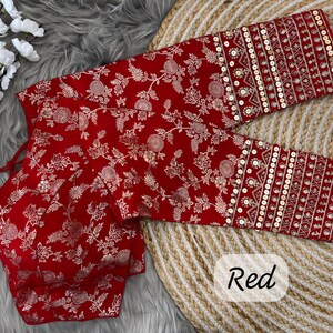 May include: A red blouse featuring a white floral pattern and long sleeves with gold sequin embellishments. The garment includes a tie-up detail at the neckline and a structured bodice, showcasing intricate embroidery throughout the fabric design.
