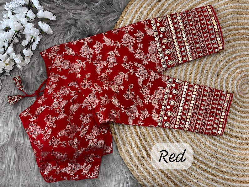 May include: A red blouse featuring a white floral pattern and long sleeves with gold sequin embellishments. The garment includes a tie-up detail at the neckline and a structured bodice, showcasing intricate embroidery throughout the fabric design.