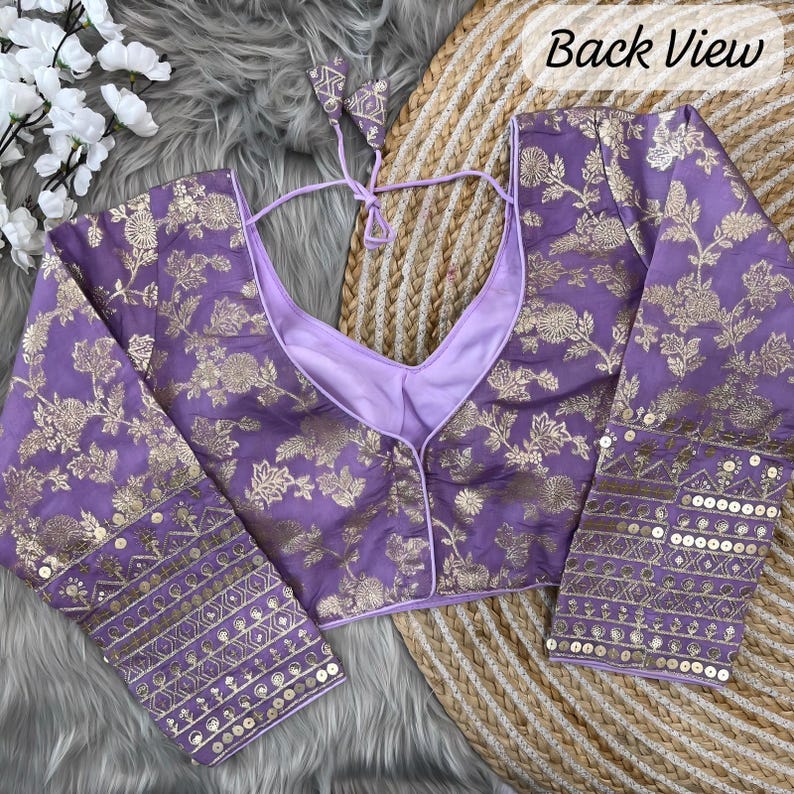 May include: A lavender blouse featuring a gold floral pattern, long sleeves, and a deep V-neckline with tie-up back closures. The cuffs are embellished with intricate gold embroidery and sequins. The text Back View is visible in the corner.