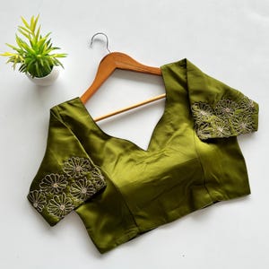 Puede incluir: Blusa verde oliva con cuello en V y mangas cortas. Las mangas están adornadas con patrones florales bordados en un color contrastante. La blusa está colgada en una percha de madera sobre un fondo blanco.