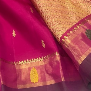 May include: A vibrant fuchsia silk saree featuring a wide gold zari border with intricate woven patterns. The fabric is adorned with small gold leaf motifs scattered across the main body, complemented by a deep purple hemline and metallic gold accents.