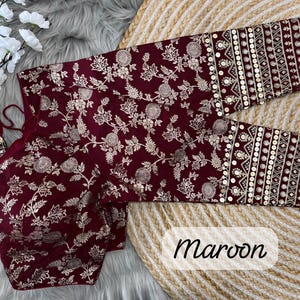 May include: A maroon blouse featuring a gold floral pattern throughout the fabric. The sleeves are adorned with horizontal bands of intricate gold embroidery and circular sequins. The garment includes thin maroon ties with decorative tassels.