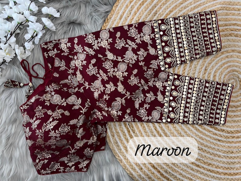 May include: A maroon blouse featuring a gold floral pattern throughout the fabric. The sleeves are adorned with horizontal bands of intricate gold embroidery and circular sequins. The garment includes thin maroon ties with decorative tassels.