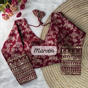 May include: A maroon blouse featuring a deep V-neckline and long sleeves with intricate gold floral patterns. The cuffs are embellished with horizontal bands of gold sequins and embroidery. A matching fabric tie is attached at the neckline.