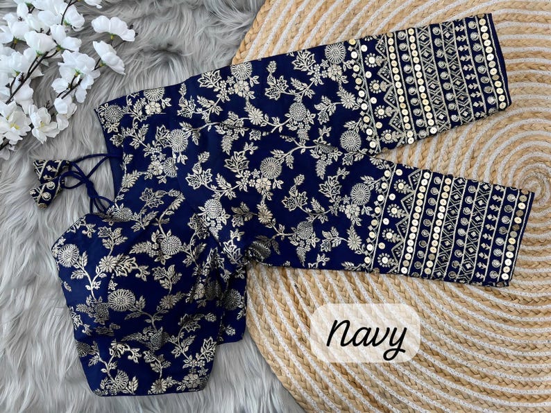 May include: A navy blue blouse featuring a gold floral pattern and long sleeves with horizontal bands of gold embroidery and sequins. The garment includes a tie-back closure with tassels and is labeled with the word navy in the bottom corner.