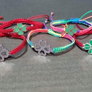 Op de afbeelding: Vijf rode en veelkleurige touw armbanden met een klavertje vier bedel. Elke bedel is van een andere kleur, waaronder groen, zilver en rood.