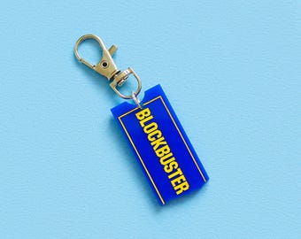 May include: A blue rectangular keychain with the word "BLOCKBUSTER" in yellow lettering. The keychain has a gold-colored clasp. The background is a solid light blue.