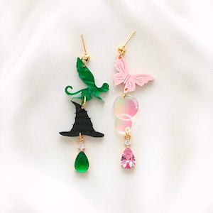May include: A pair of mismatched earrings. One earring features a black witch hat, a green gem, and a green bat. The other earring has a pink butterfly, iridescent circles, and a pink gem. Both earrings have gold-colored posts.
