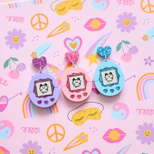 May include: Three colorful earrings in the shape of retro handheld game consoles. Each earring features a pixelated character on the screen and a glittery heart at the top. The earrings are purple, pink, and blue.