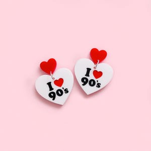 May include: A pair of heart-shaped earrings. Each earring features a white heart with the text "I ❤️ 90's" in black, and a small red heart. A red heart tops each earring. The earrings are displayed on a pink background.