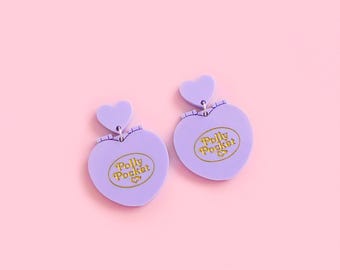 Peut inclure: Une paire de boucles d'oreilles en forme de cœur lilas, avec un petit cœur en haut. Chaque boucle d'oreille porte le logo "Polly Pocket" en or. Les boucles d'oreilles sont posées sur un fond rose clair.