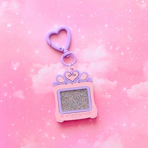 May include: A pink and purple keychain resembling a retro television, featuring a heart-shaped clasp. The screen is filled with glitter. The top of the TV has a crown design. The background is pink with sparkles.