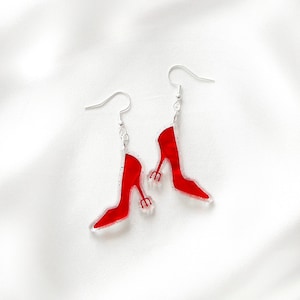 May include: A pair of red high heel earrings with silver hooks. The earrings are shaped like high heels with a three-pronged design on the heel. The earrings are set against a white background.
