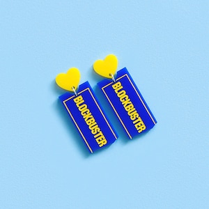 May include: A pair of blue and yellow Blockbuster earrings. Each earring features a yellow heart-shaped top and a rectangular blue pendant with the word "BLOCKBUSTER" in yellow. The earrings are set against a light blue background.
